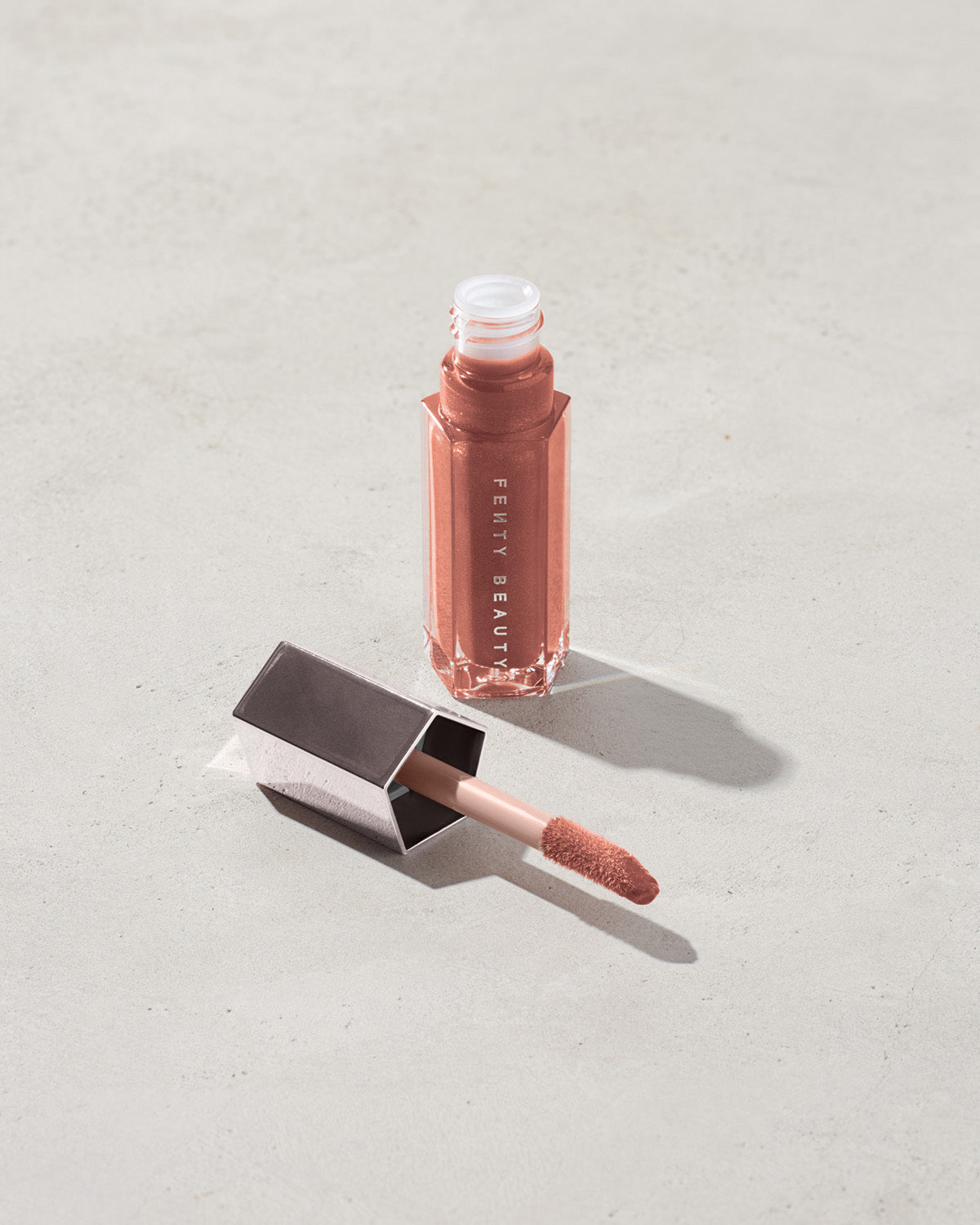 An open tube of Gloss Bomb Universal Lip Luminizer in the shade Fenty Glow on a grey background.