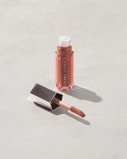An open tube of Gloss Bomb Universal Lip Luminizer in the shade Fenty Glow on a grey background.