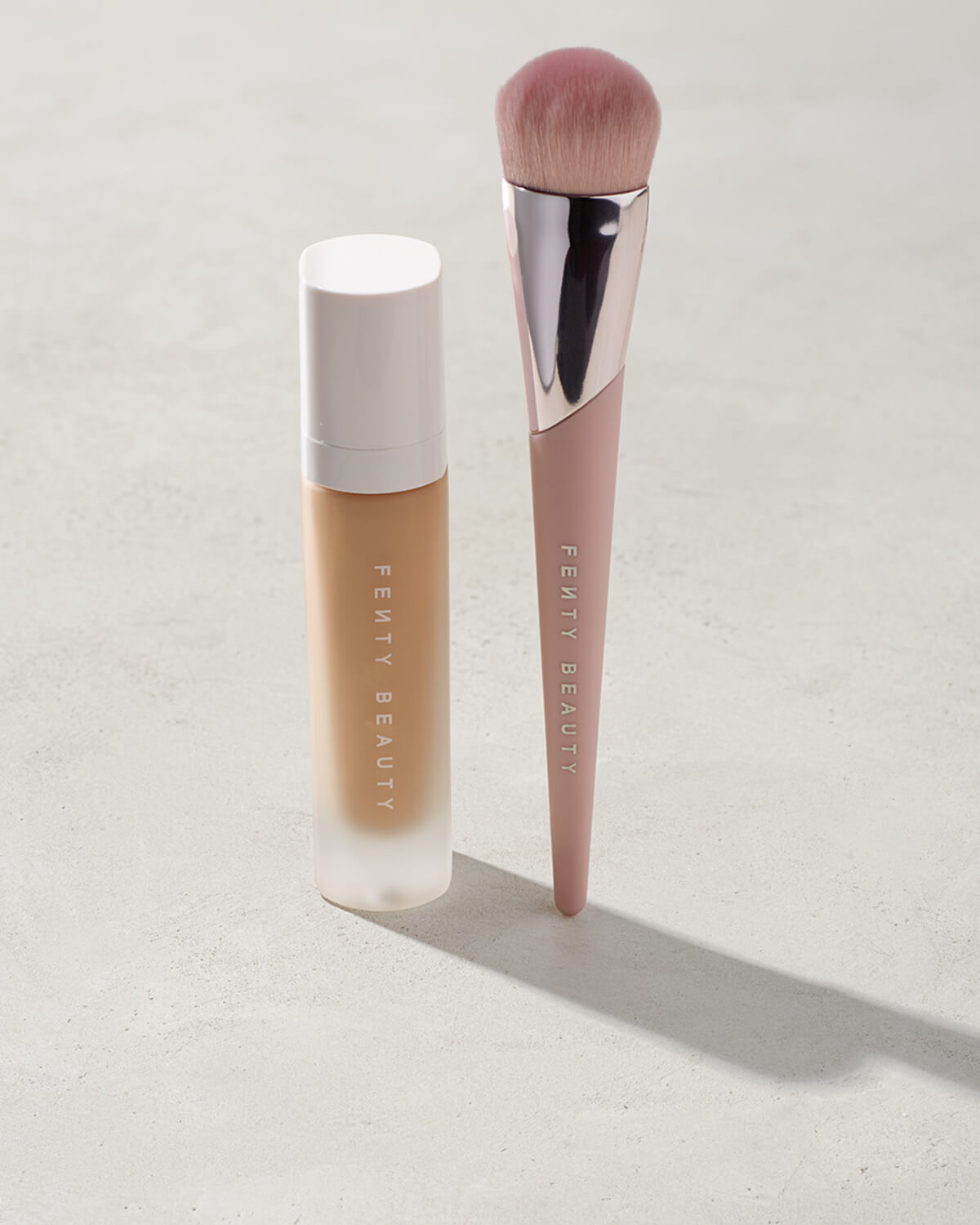Full-Bodied Foundation Brush 110 standing upright next to a Fenty Beauty liquid foundation on a concrete backdrop.