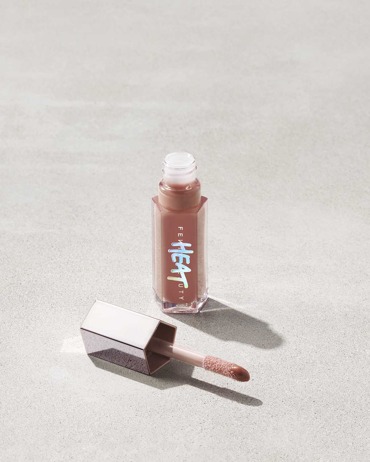 Fenty Beauty Gloss Bomb Heat Universal Lip Luminizer + Plumper in shade Fenty Glow Heat open showing doe foot applicator on concrete background