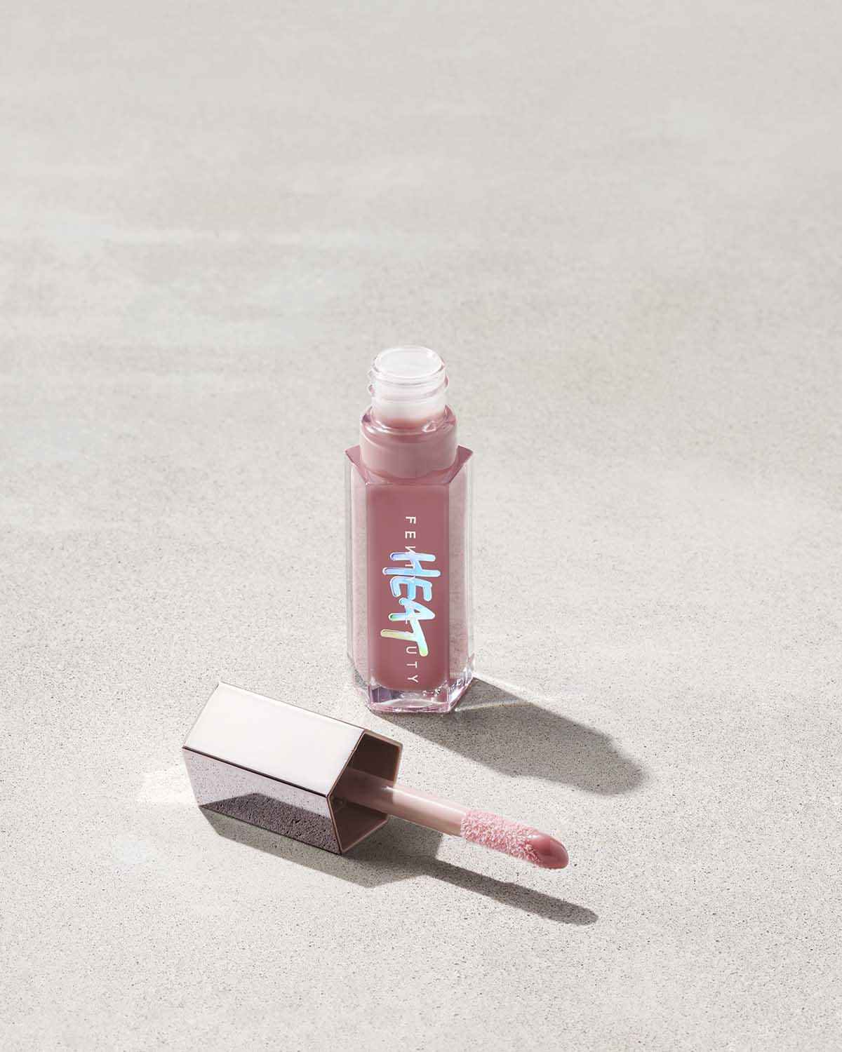 open tube of Fenty Beauty Gloss Bomb Heat Universal Lip Luminizer + Plumper in shade Fussy Heat with doe foot applicator on concrete background