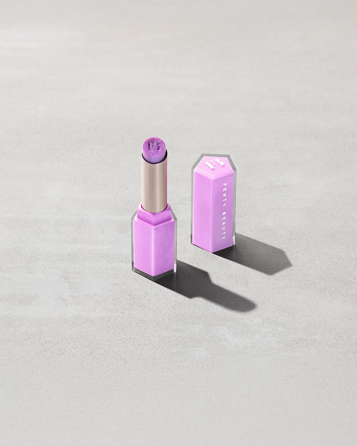 Open product shot of Gloss Bomb Stix Shimmer in shade Ice Amethyst on a concrete background.