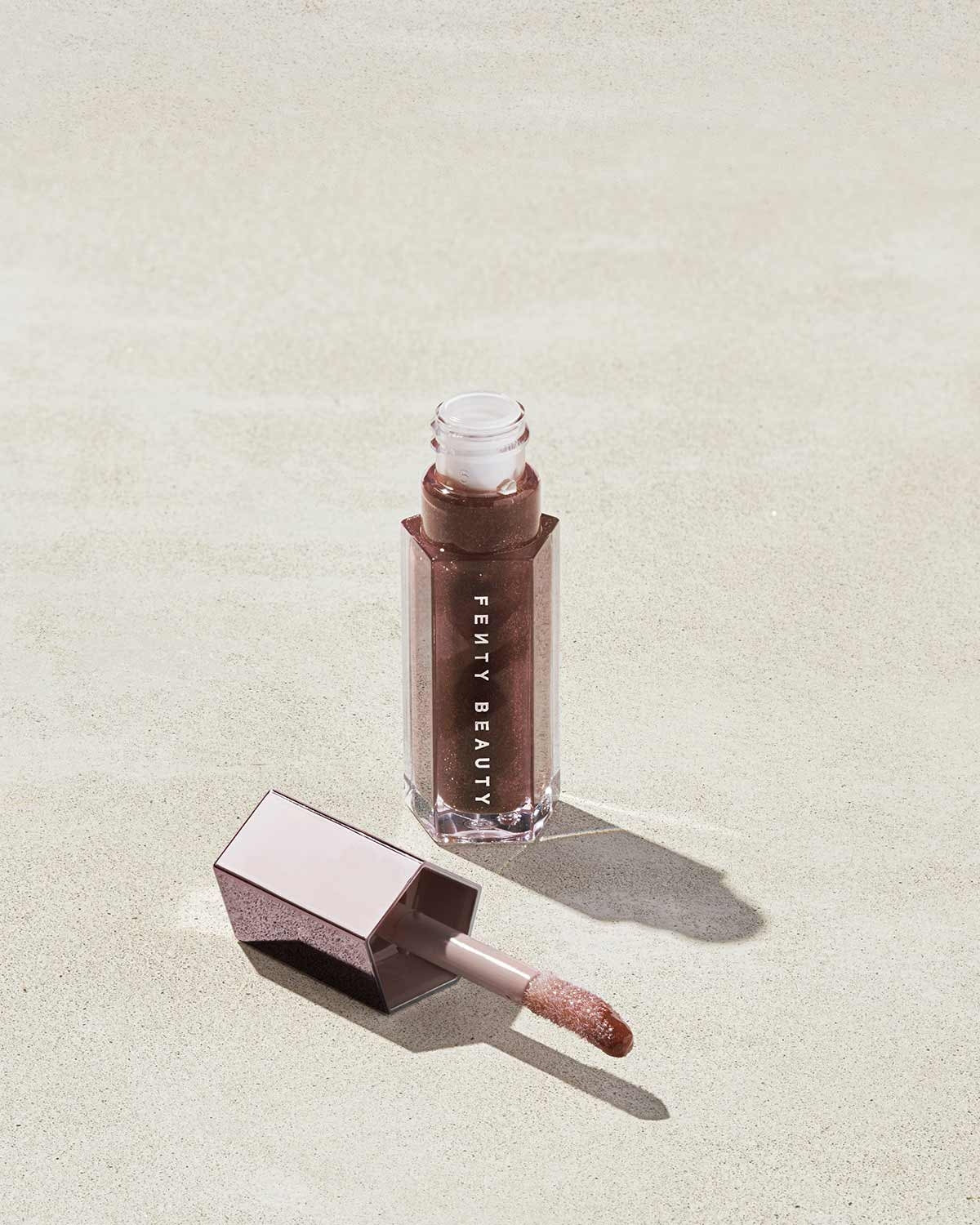 An open tube of Gloss Bomb Universal Lip Luminizer in the shade Hot Chocolit on a grey background.