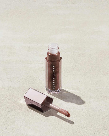 An open tube of Gloss Bomb Universal Lip Luminizer in the shade Hot Chocolit on a grey background.