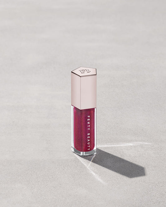 A closed tube of Gloss Bomb Universal Lip Luminizer in the shade Fuschia Flex on a grey background.